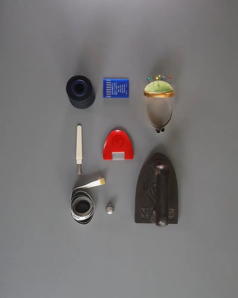 Outils de couture disposés sur fond gris : fil, aiguilles, pique-épingles, craie, découd-vite, mètre, dé et fer ancien